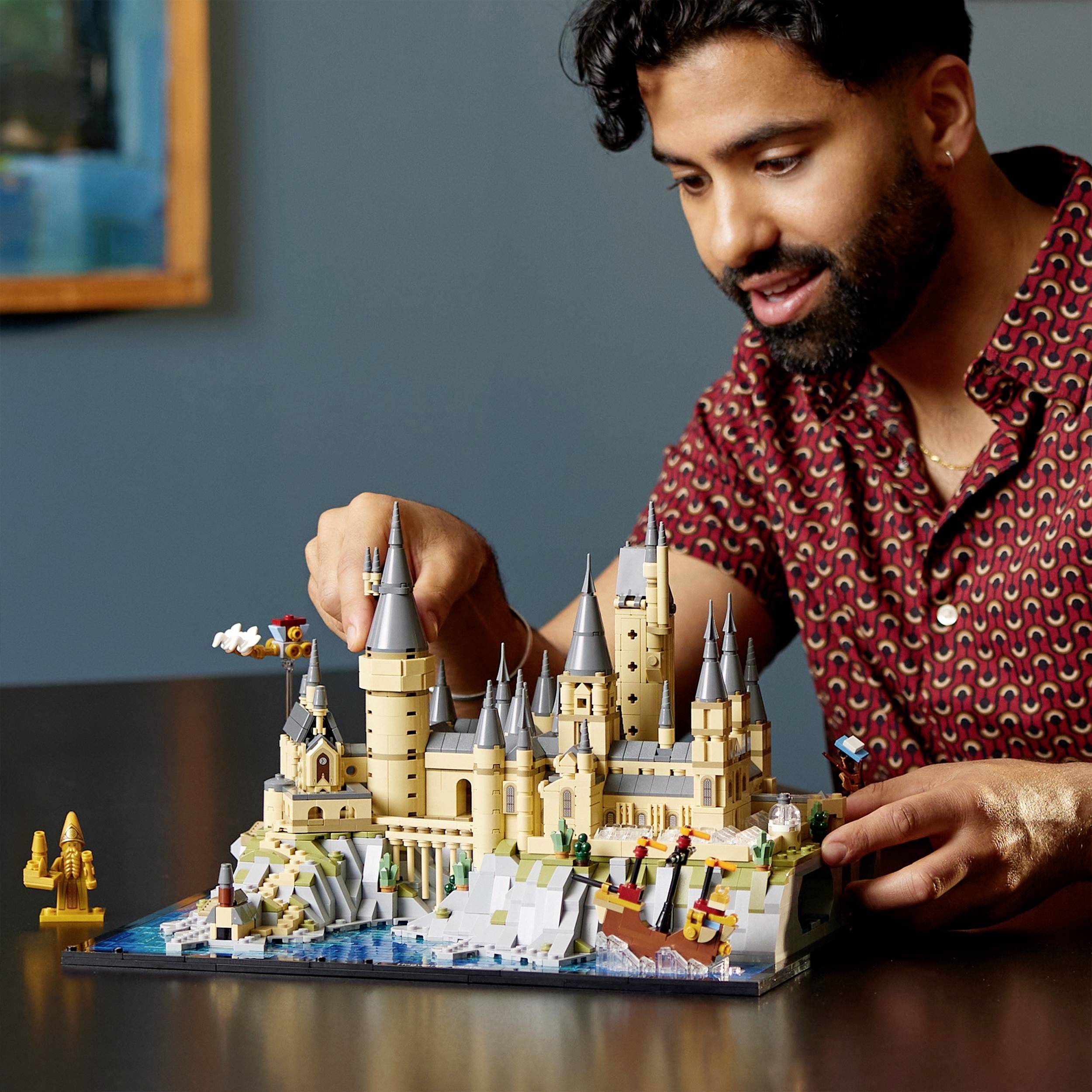 A person is building a detailed model of an imaginative castle on a table. The person is wearing a checked shirt and looks intently interested.