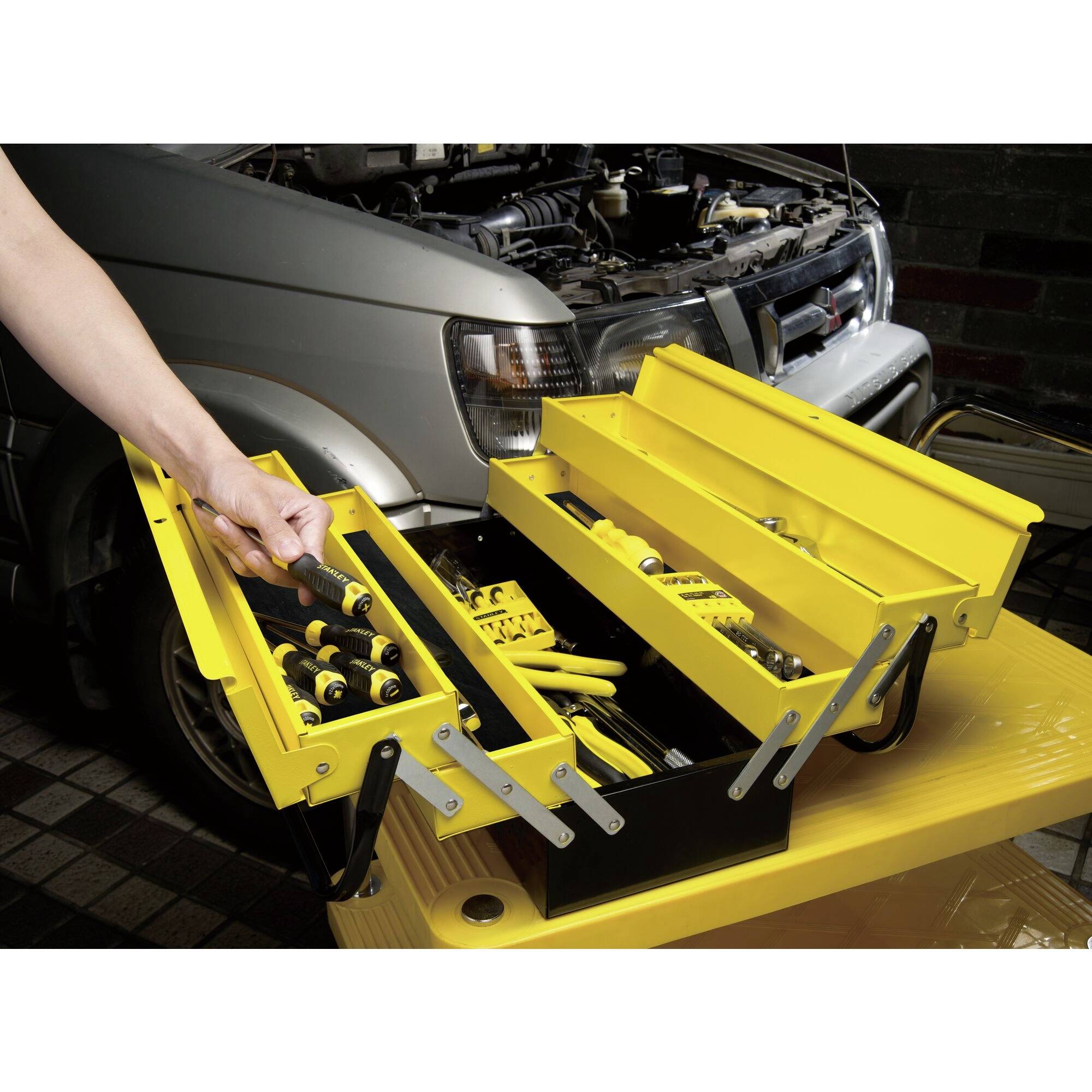A person removes a screwdriver from a yellow toolbox on a trolley in front of a car with an open bonnet.