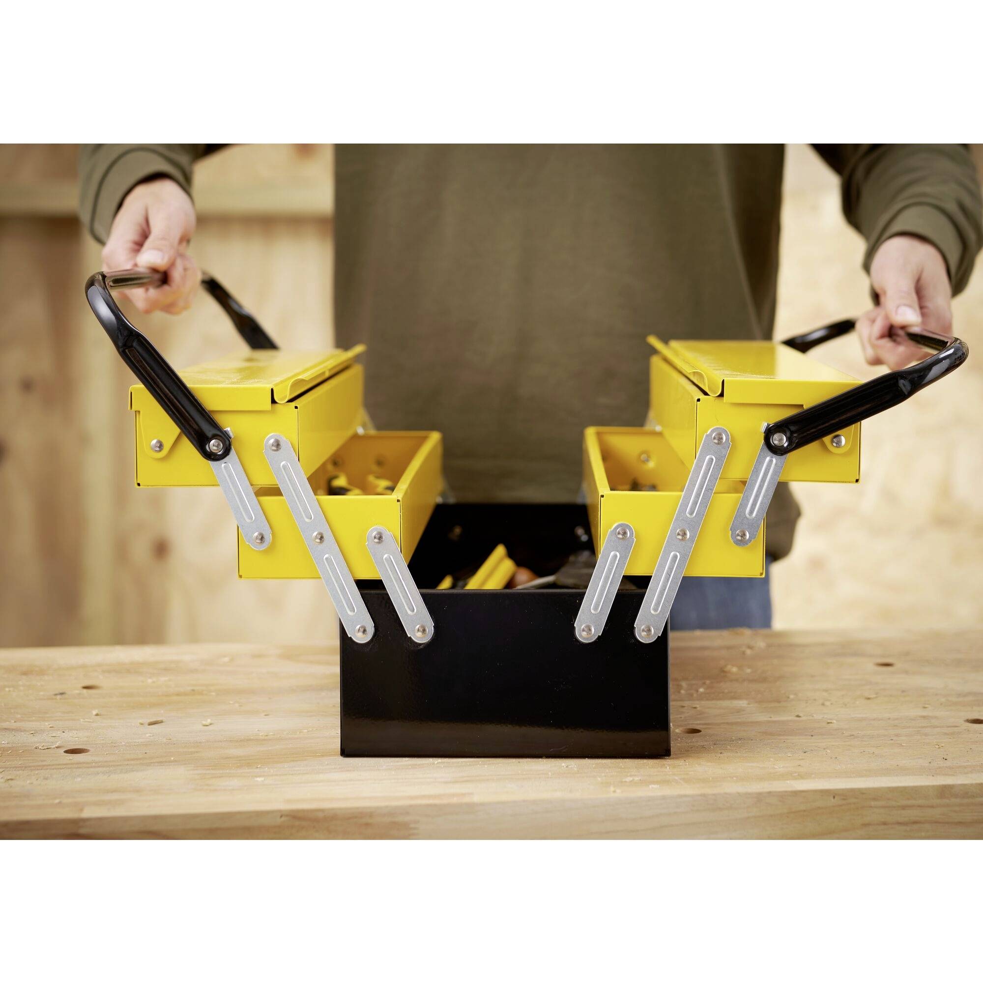 A person opens a yellow toolbox with black handles on a wooden workshop table. The box has multiple compartments.