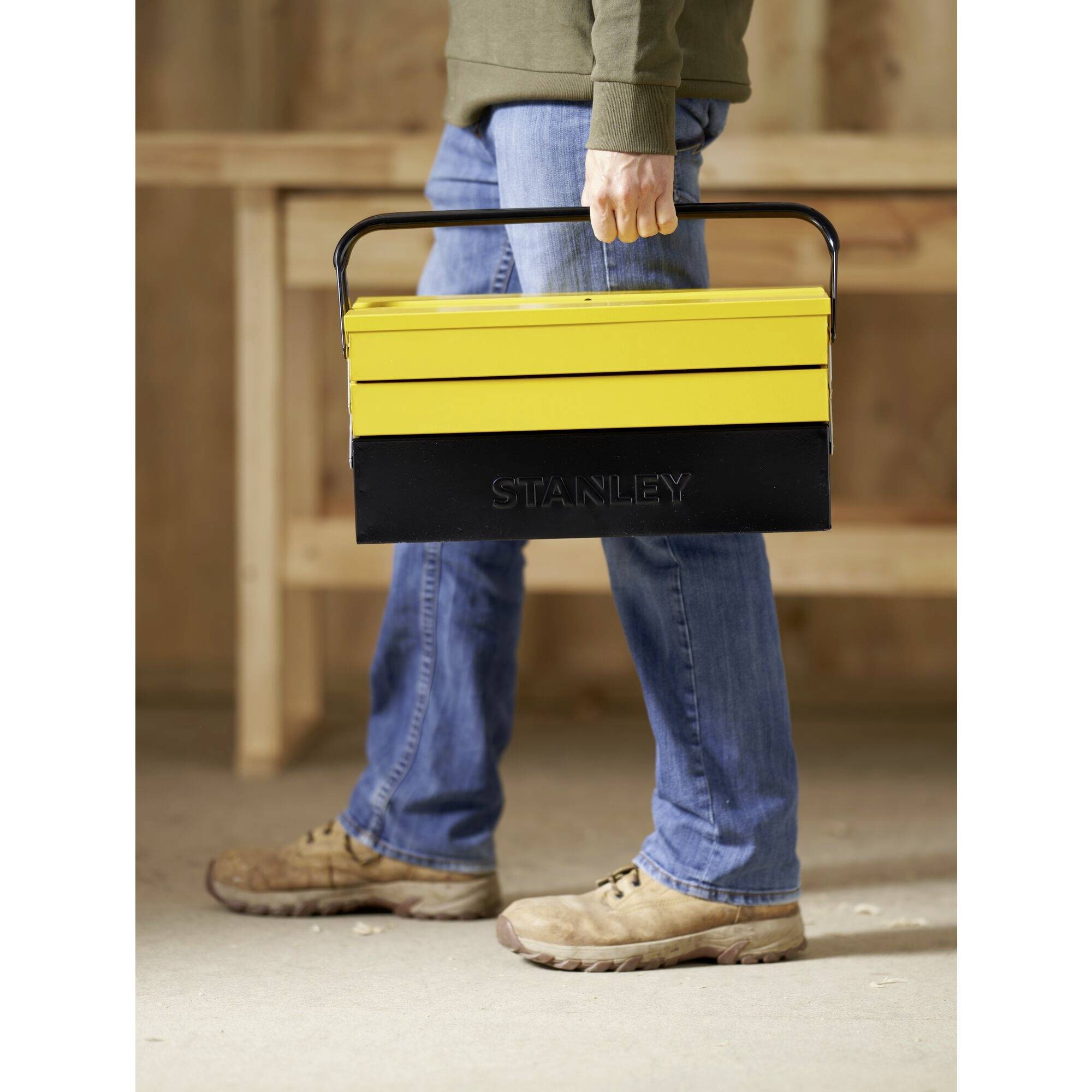 A person is carrying a yellow Stanley toolbox. The person is wearing jeans and beige boots and walking on a wooden floor.
