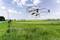 A remote-controlled helicopter flies over a green field with a red and white farmhouse in the background beneath a cloudy sky.