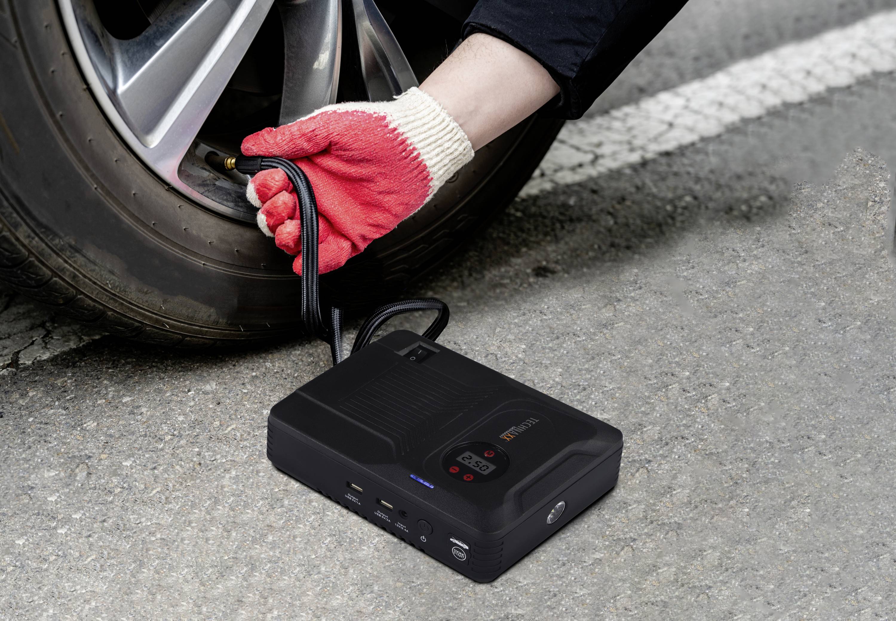 A hand wearing red gloves is pumping air into a car tyre with a portable compressor on a road.