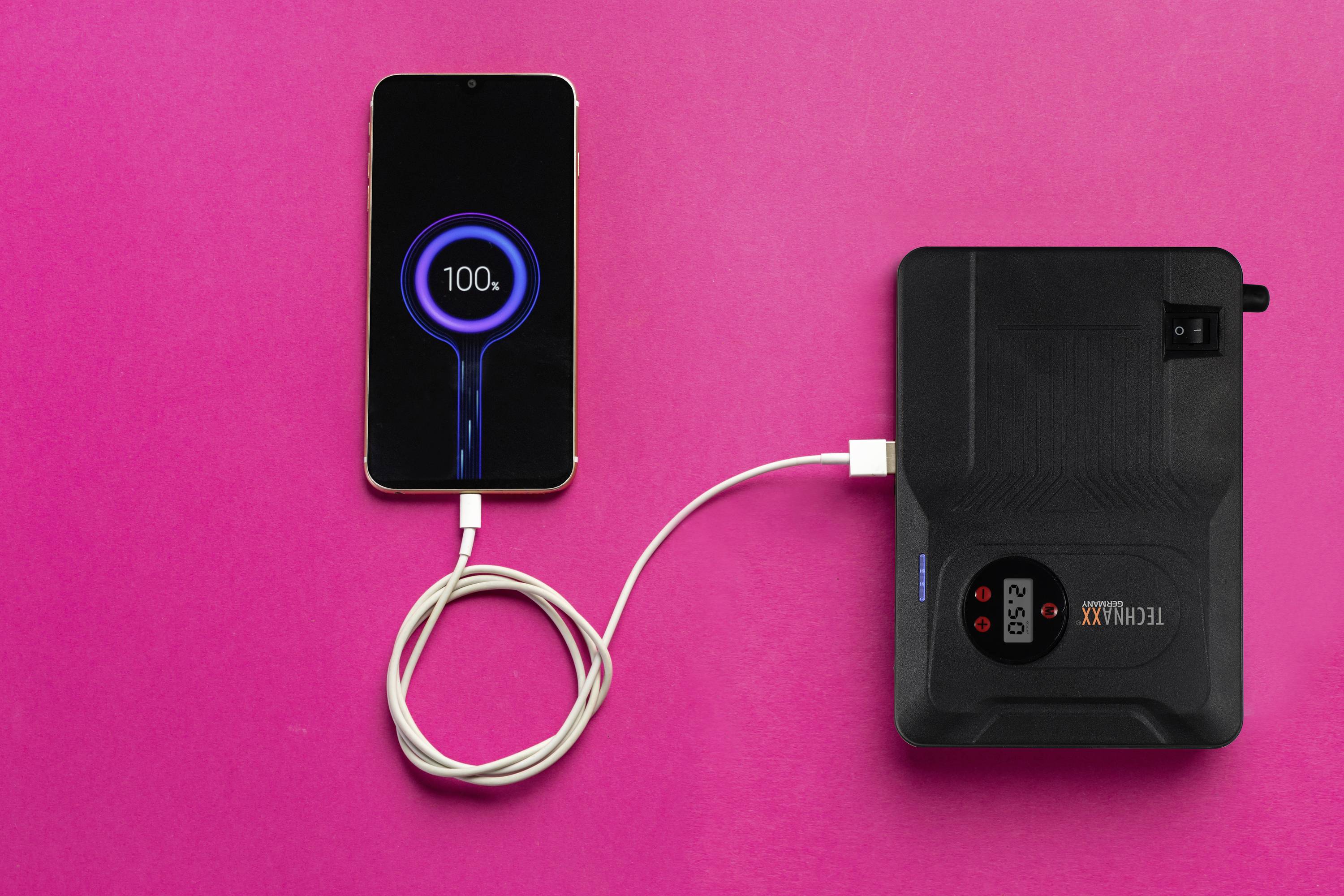 A smartphone on a pink background, connected to a power bank, showing 100% battery charge.