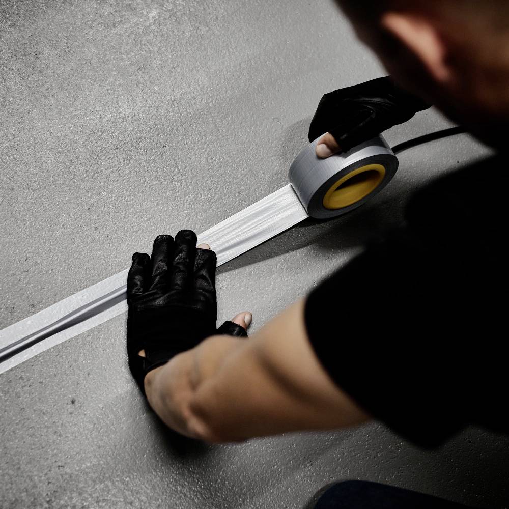 A person is wearing black gloves and sticking grey adhesive tape onto a concrete floor. The scene depicts careful workmanship.