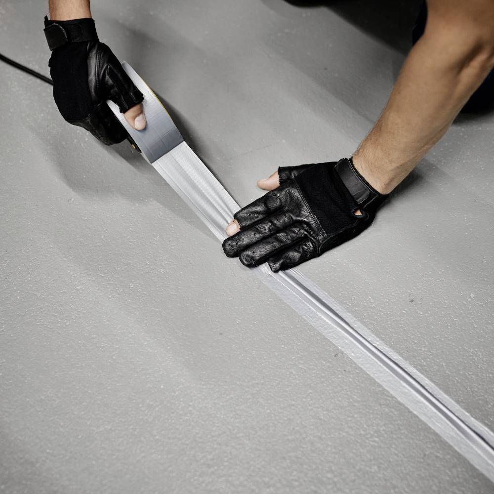 A person wearing gloves is cutting silver duct tape on a grey floor.