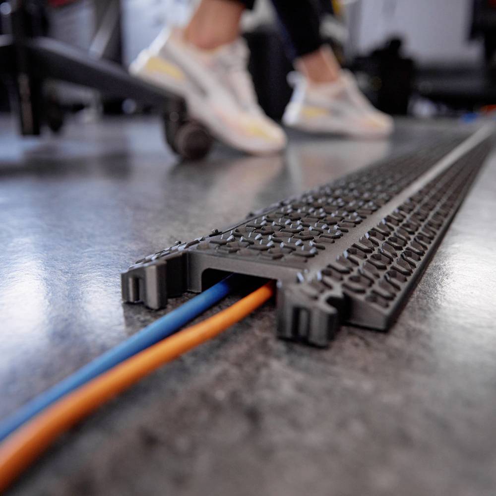 Cable protection on the floor with blue and orange cables in an office. A person wearing trainers is blurred in the background.