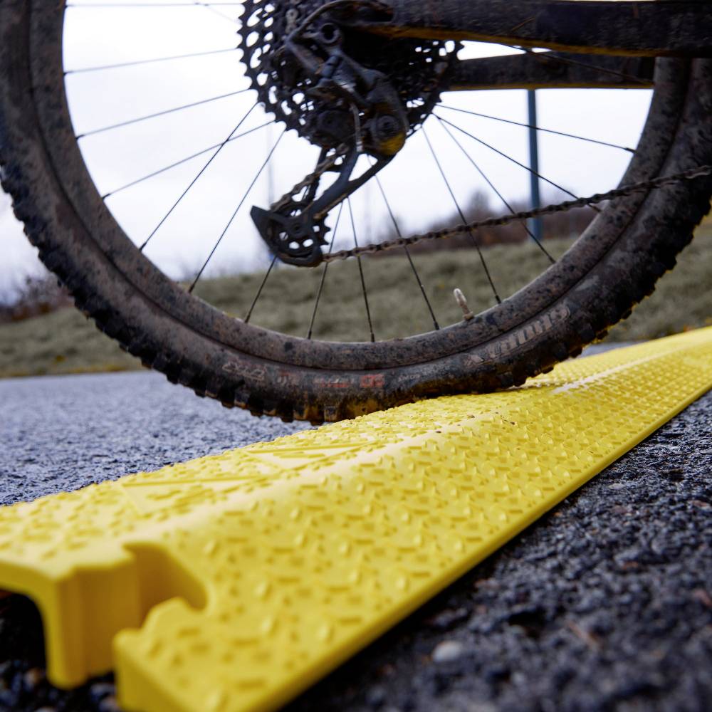 Muddy bicycle tyre crosses yellow, ribbed plastic mat on rocky ground, likely for improved grip or protection.
