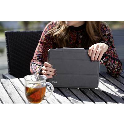 A person is sitting outside at a table, holding a closed tablet in a black case. A glass of tea is on the table.