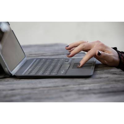 A person is typing on a laptop on a wooden table outdoors. Hands in focus, grey plastic casing, tranquil surroundings.