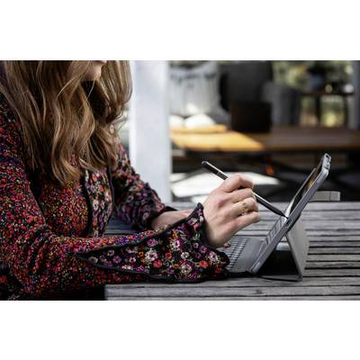 A person in a colourful blazer is writing with a pen on a tablet, which is resting on a wooden table. Blurred furniture is visible in the background.