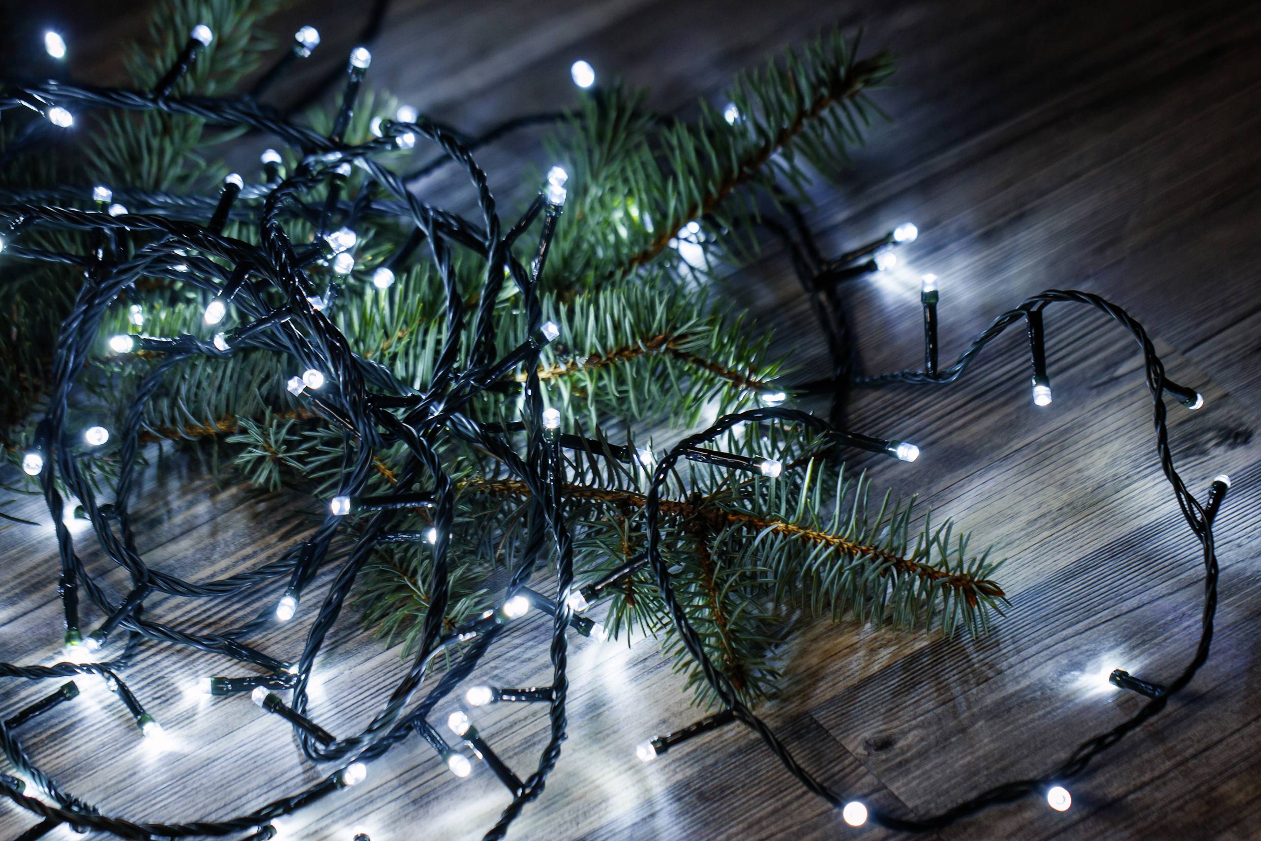 Fairy lights with white bulbs intertwined around green pine branches on a wooden floor, creating a festive decoration in a cosy room.
