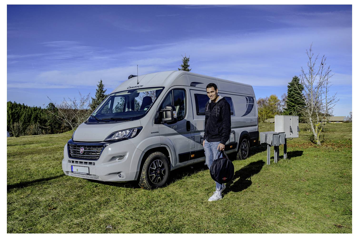 Man stands beside a silver camper van parked on a grassy field under a blue sky.