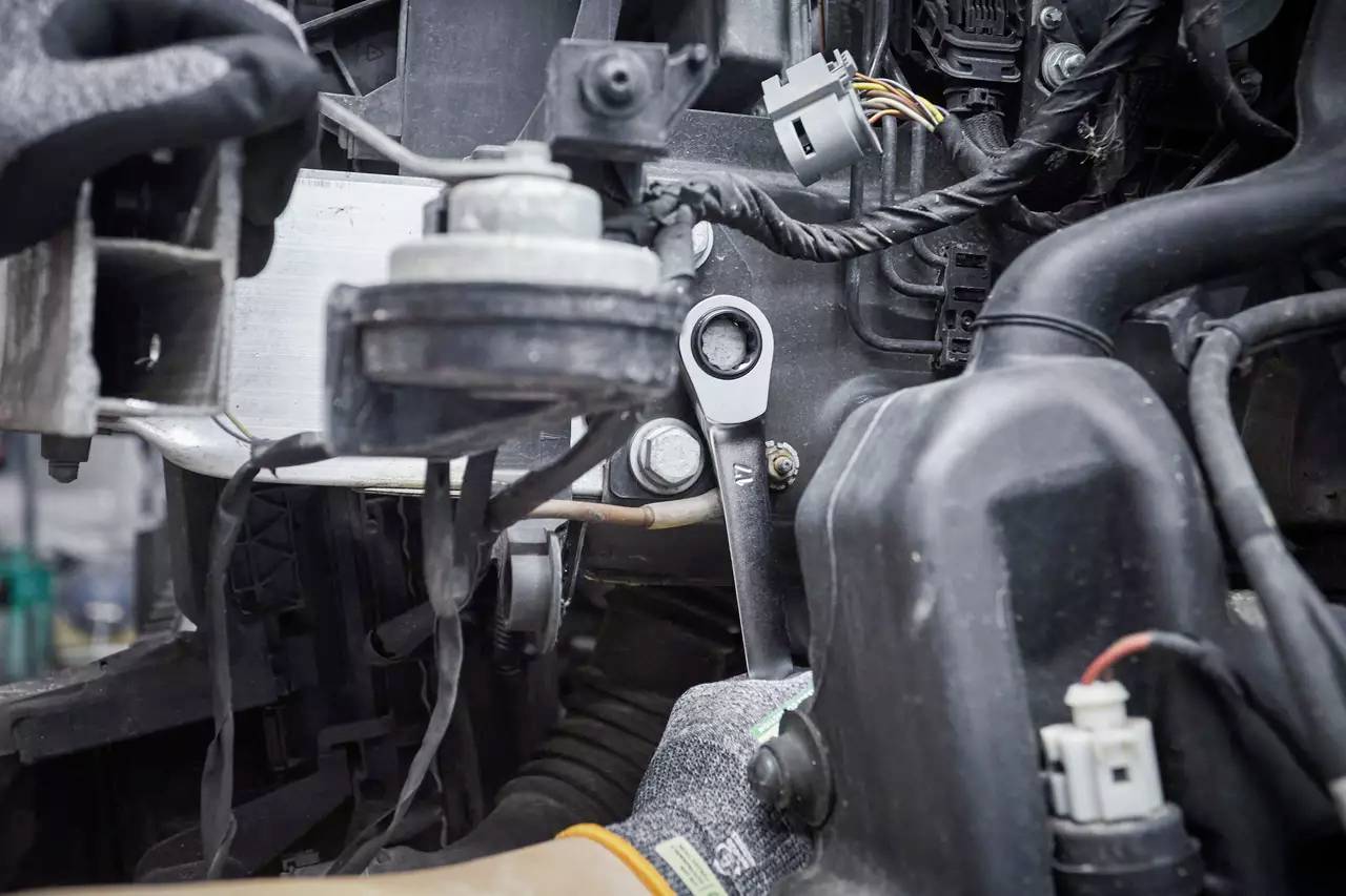 A mechanic is working in the engine bay of a car with a spanner. Various cables and components are visible.