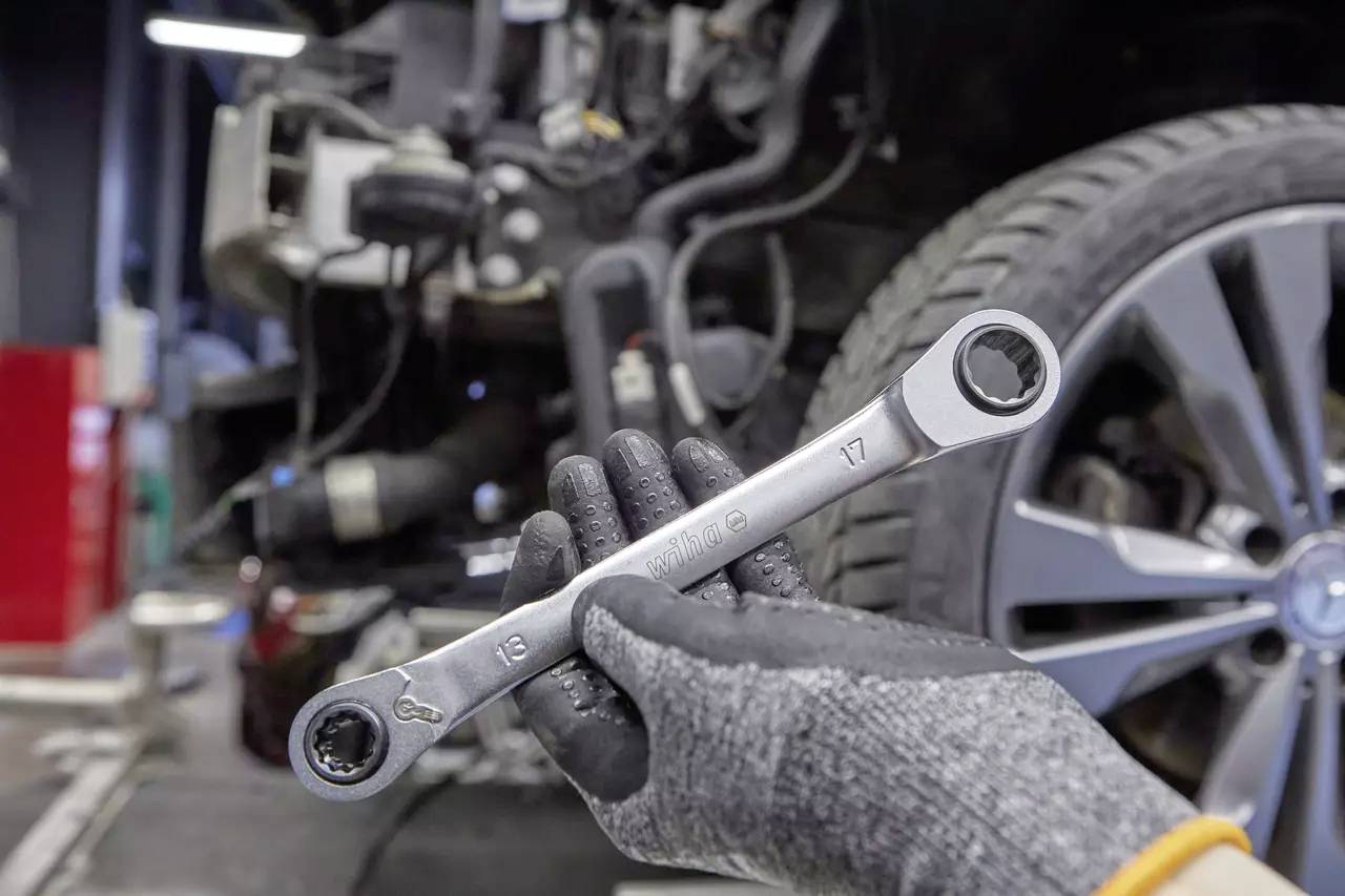 A mechanic is holding a socket wrench in front of a car in a workshop. The front tyre is visible, with the focus on the tool.