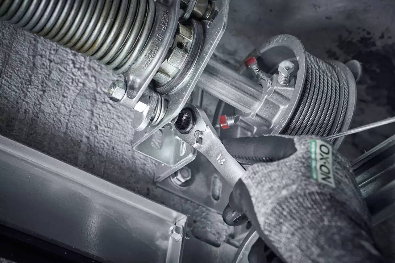 A hand wearing a glove holds a spanner and tightens a screw on a spring mechanism. Mechanical details are visible.