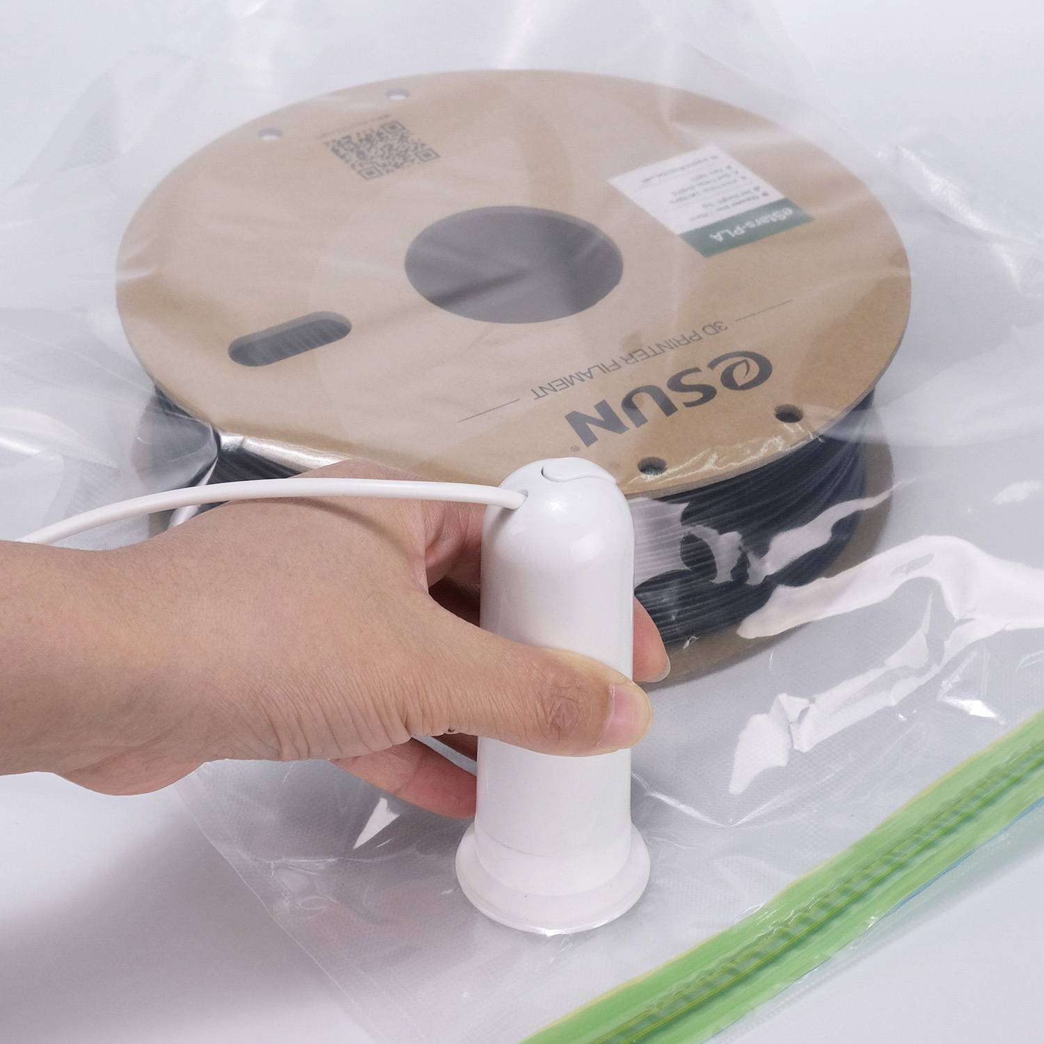 A person is compressing a plastic bag with a handheld vacuum device. Inside the bag is a spool with black filament.
