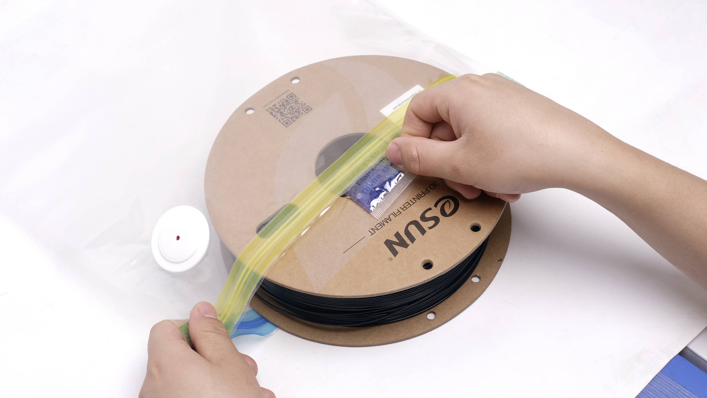 Two hands seal a plastic bag containing a roll of black filament. The filament reel is resting on a table.