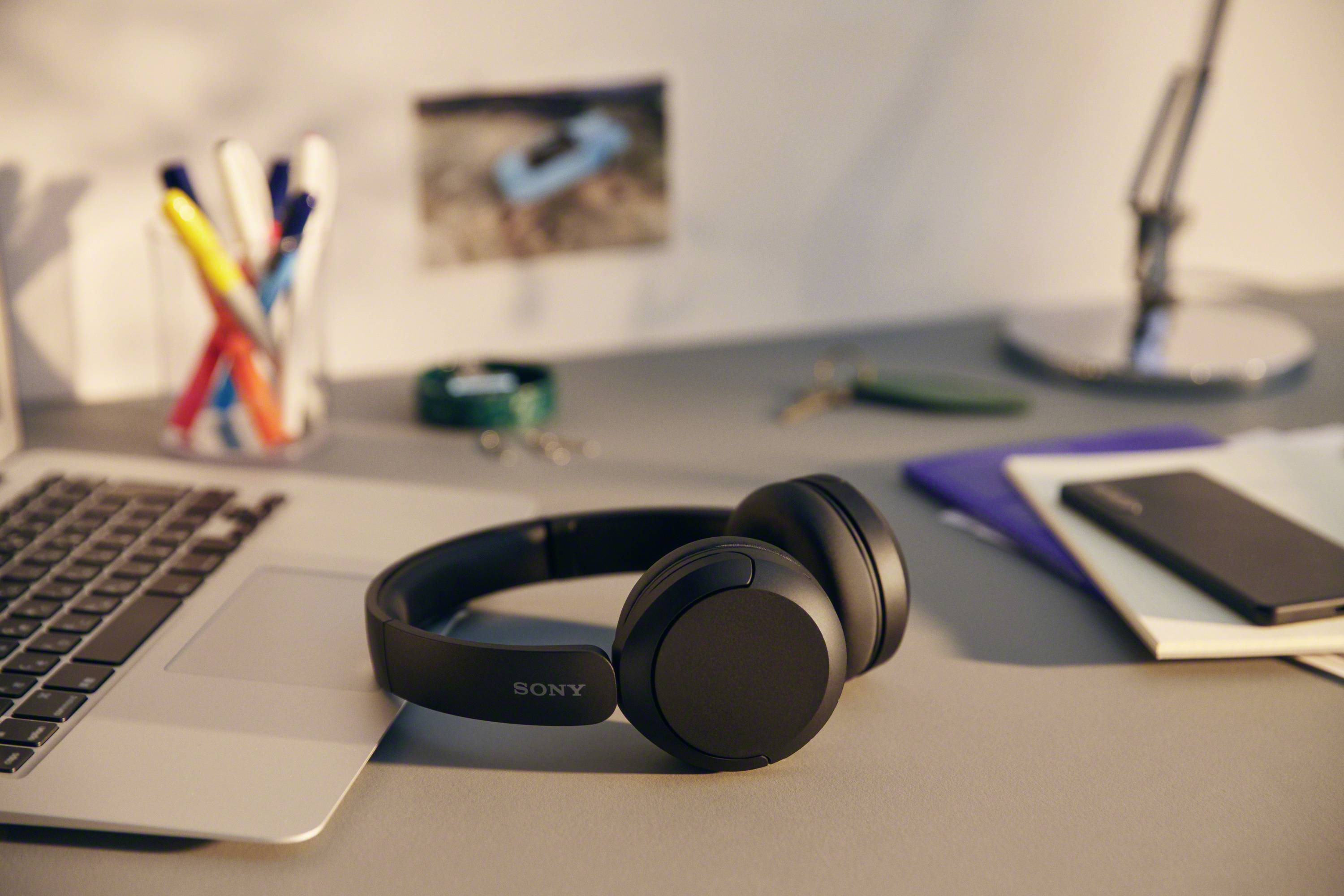 A laptop and black headphones are lying on a desk. Next to these are pens, a notepad and a lamp.