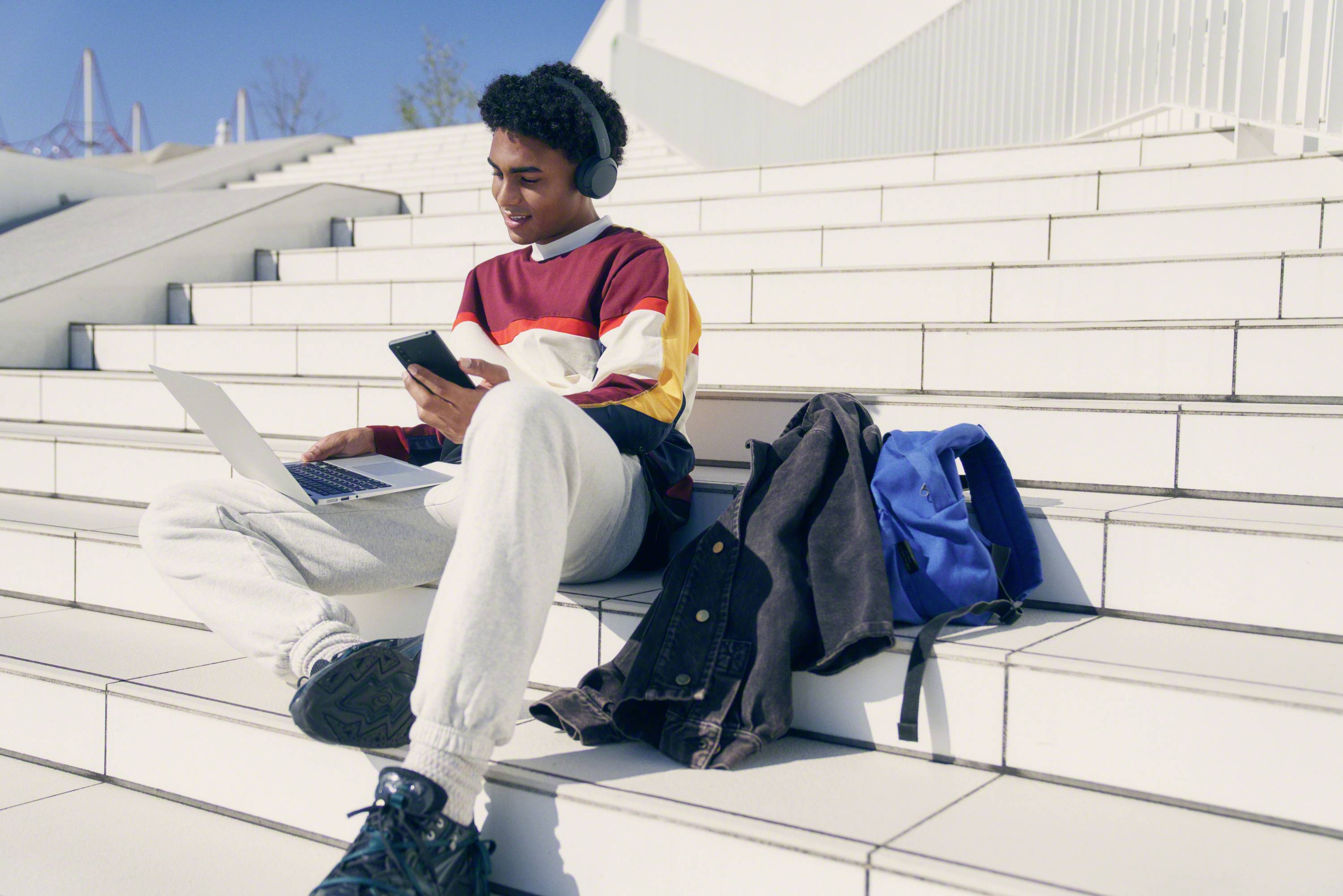 A young man is sitting on white steps, wearing headphones and using a smartphone and laptop. A jacket and a blue rucksack are lying beside him.