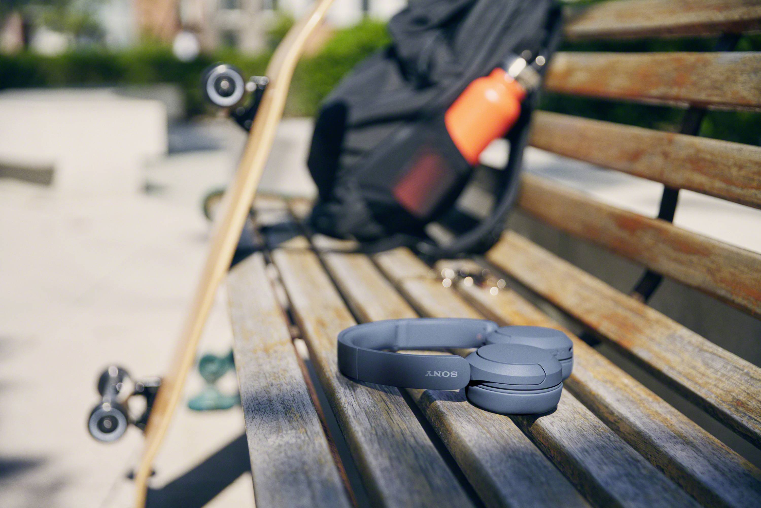 Headphones on a park bench, next to a skateboard and a black rucksack with a red water bottle. Outdoor scene.