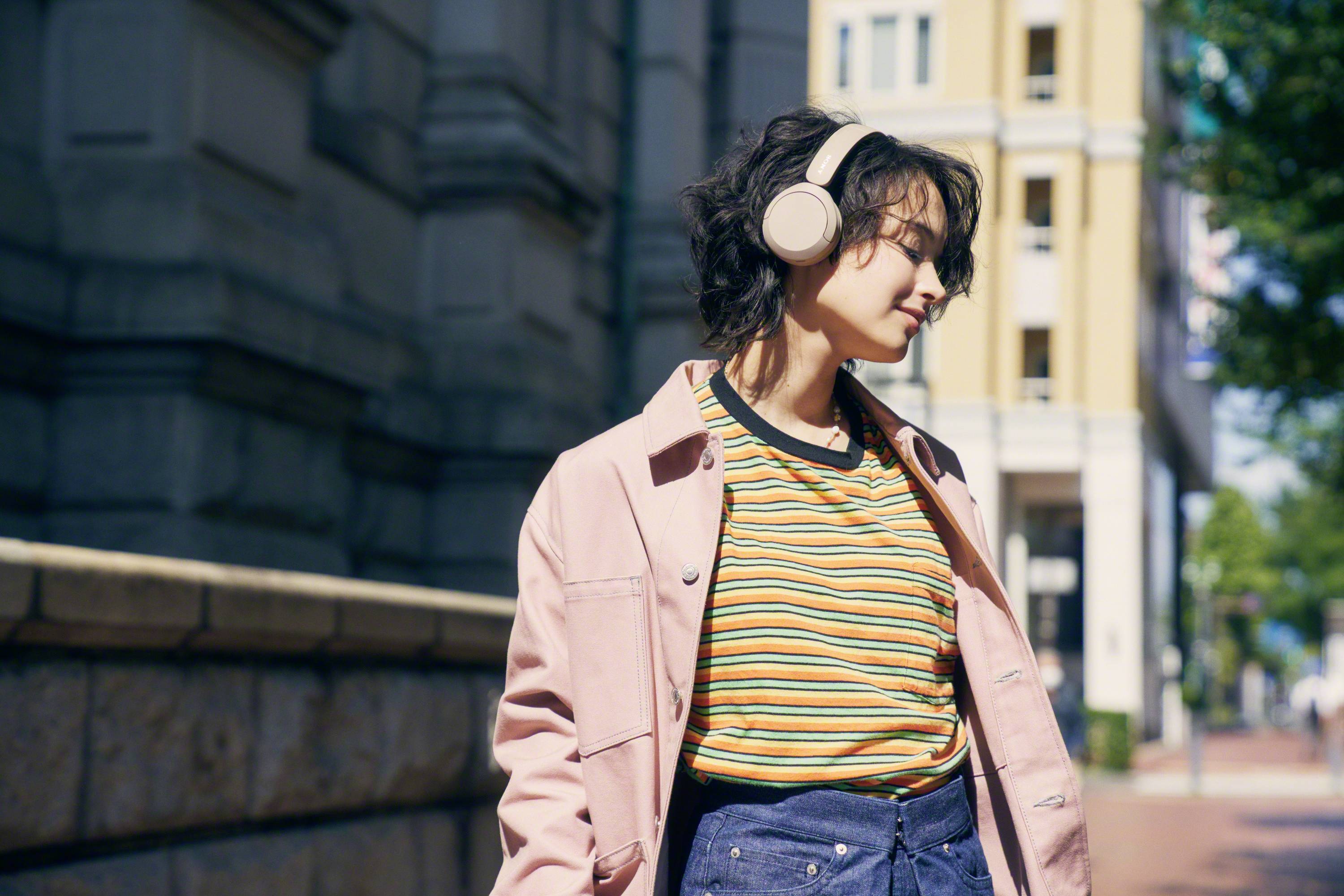 A person wearing headphones, dressed in a striped top and jacket, strolls through an urban street on a sunny day.