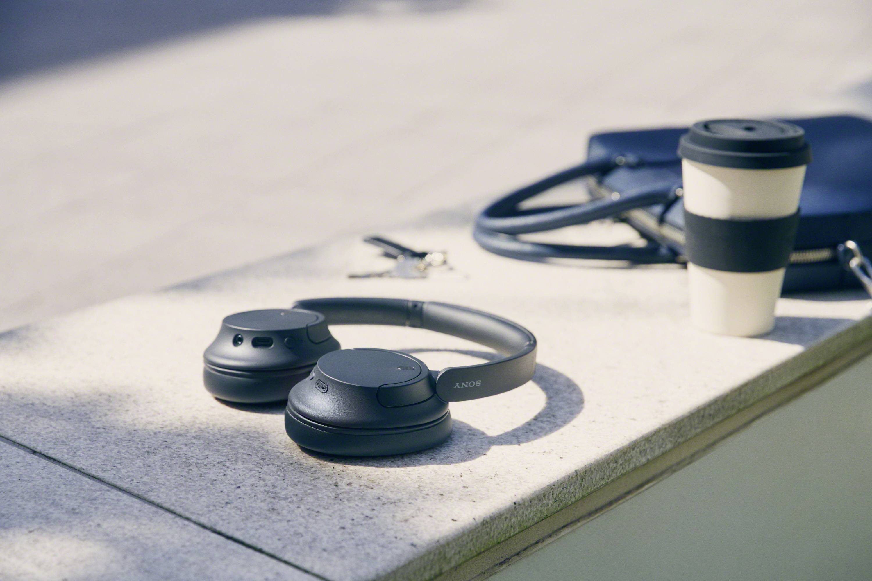 Black headphones lie on a bench next to a reusable coffee cup and a set of keys.