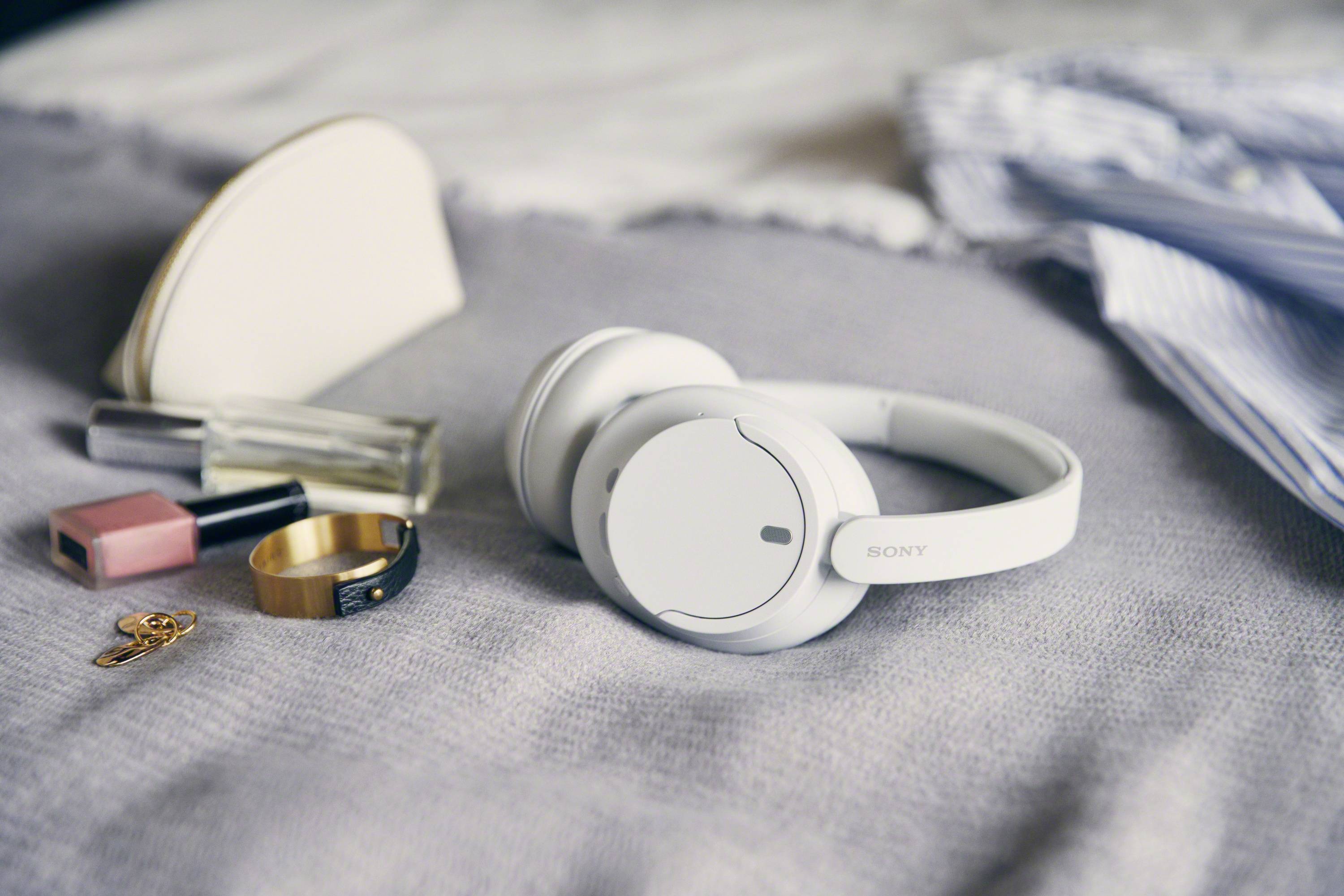 White headphones on a grey blanket, surrounded by perfume, lipstick, cream, ring and a key. An opportunity for relaxation.