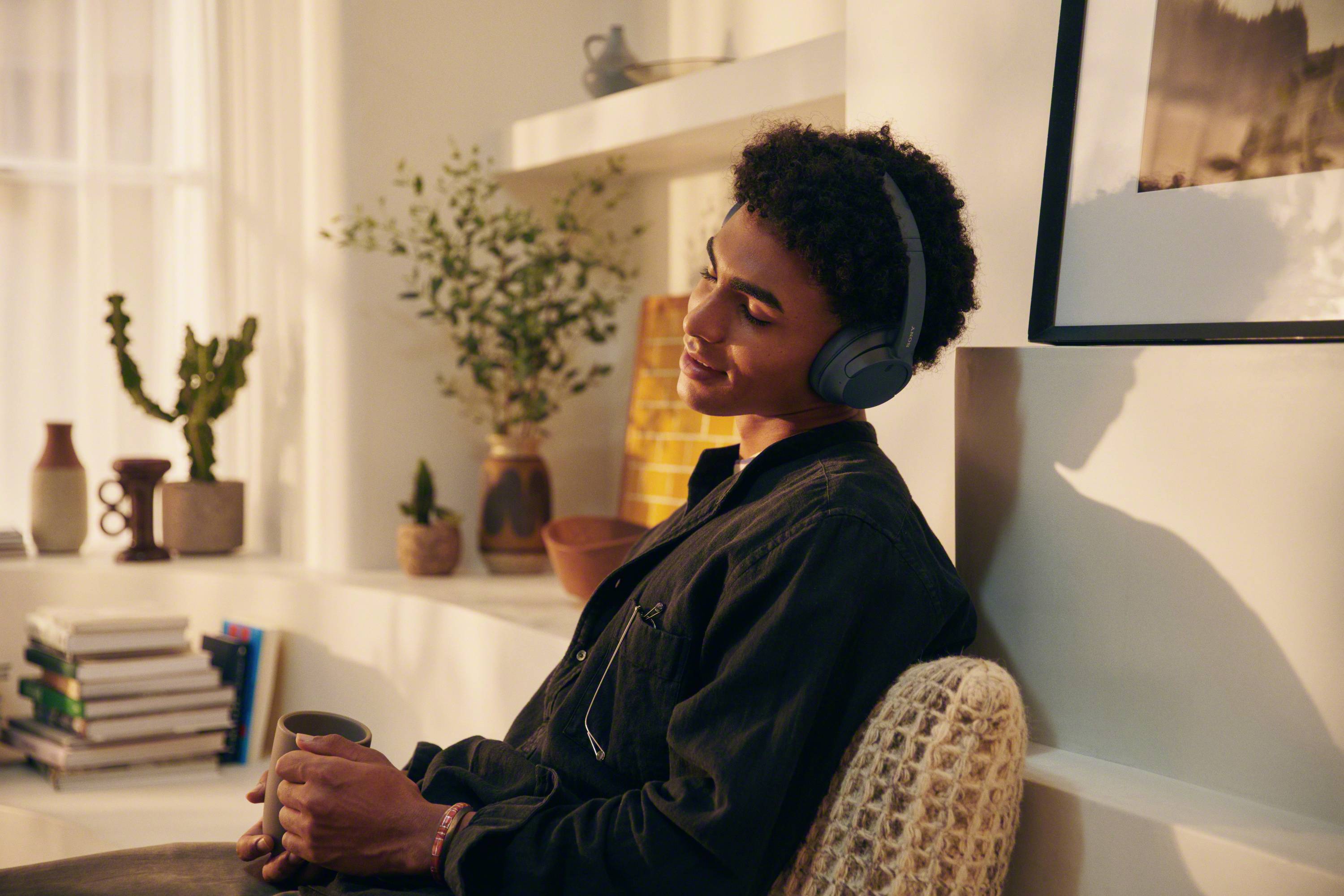 A person sits relaxed in a room, wearing headphones and holding a mug. Plants and books are visible in the background.