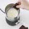 A hand holds a piece of chocolate over a milk frother, where milk is being frothed.