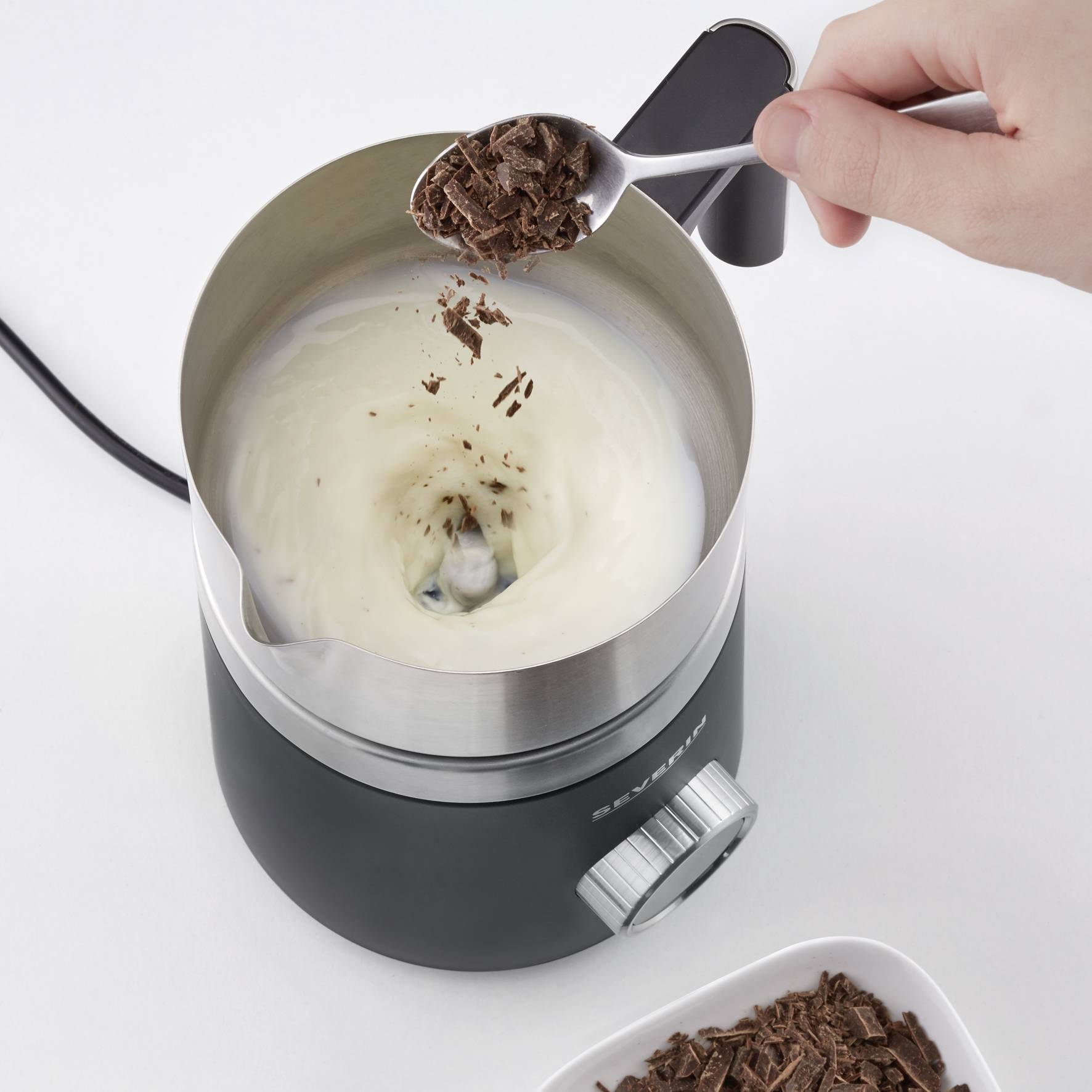 A hand sprinkles grated chocolate into a rotating milk frother, forming foam in the cup.