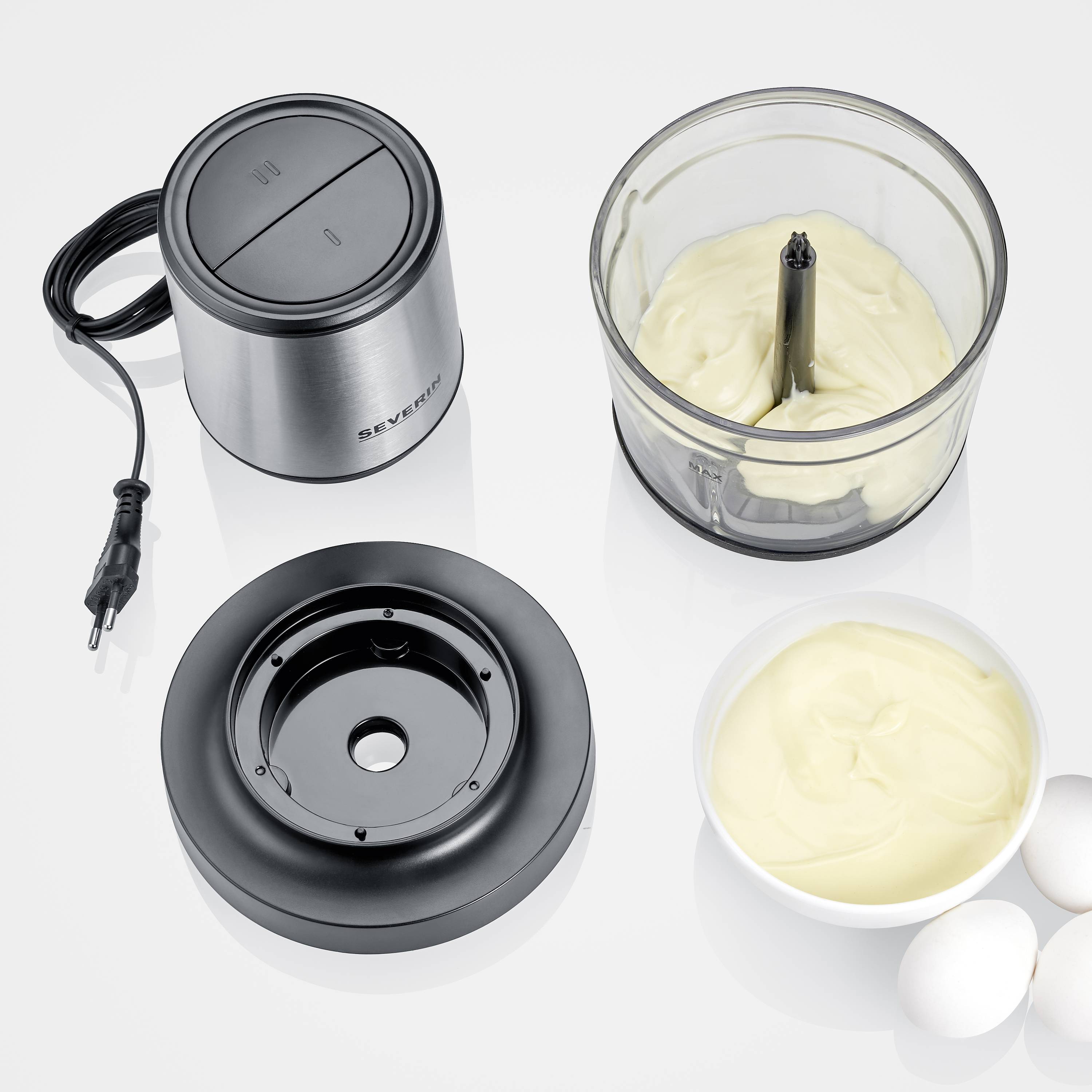 A blender with the lid removed, filled with yellow cream. Beside it, a bowl of cream and two eggs on a white surface.