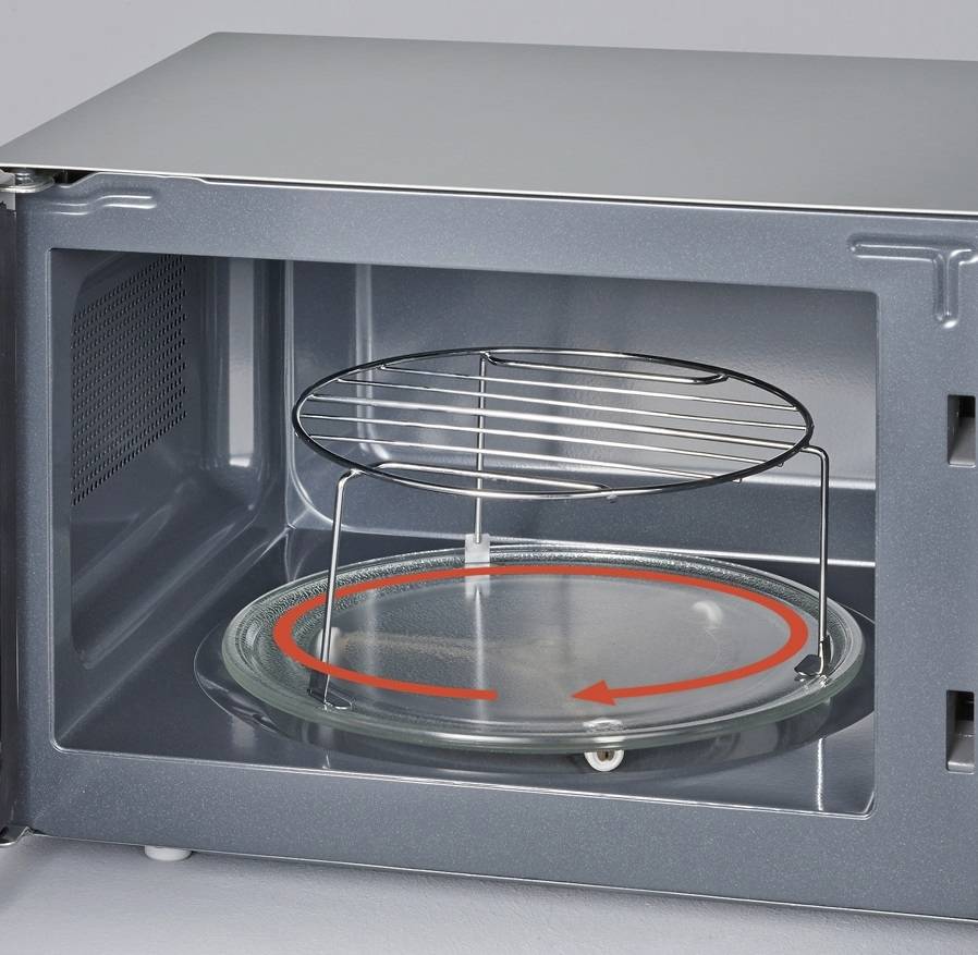 An open microwave reveals a glass turntable with a red arrow on it and a metal rack above, designed for even heating of food.