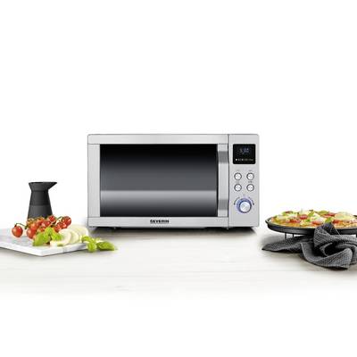 Silver microwave on kitchen table, surrounded by stacked tomatoes, onions, basil and a pizza on a plate.