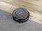 A round, black robotic vacuum cleaner cleans the floor, moving from carpet to wooden flooring. Simple operation via buttons on top.