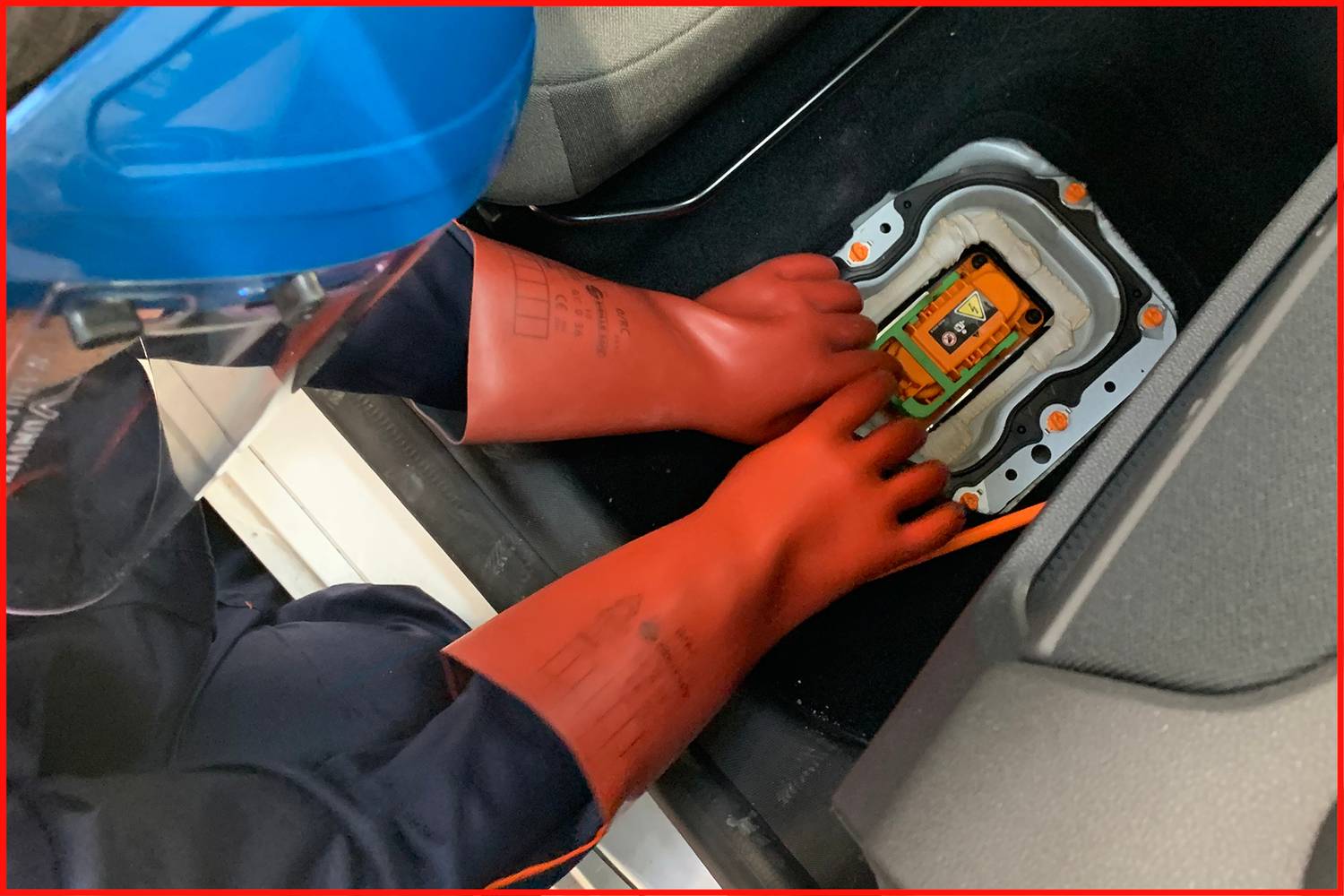 A person wearing protective clothing and gloves is working on an open battery compartment of a vehicle with orange cables.