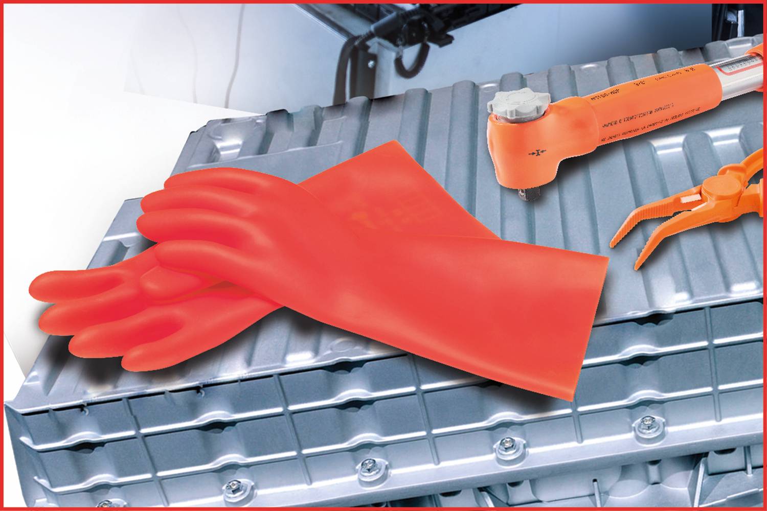 Red insulated gloves and an orange adjustable spanner lie on a silver metal casing, presumably a battery.