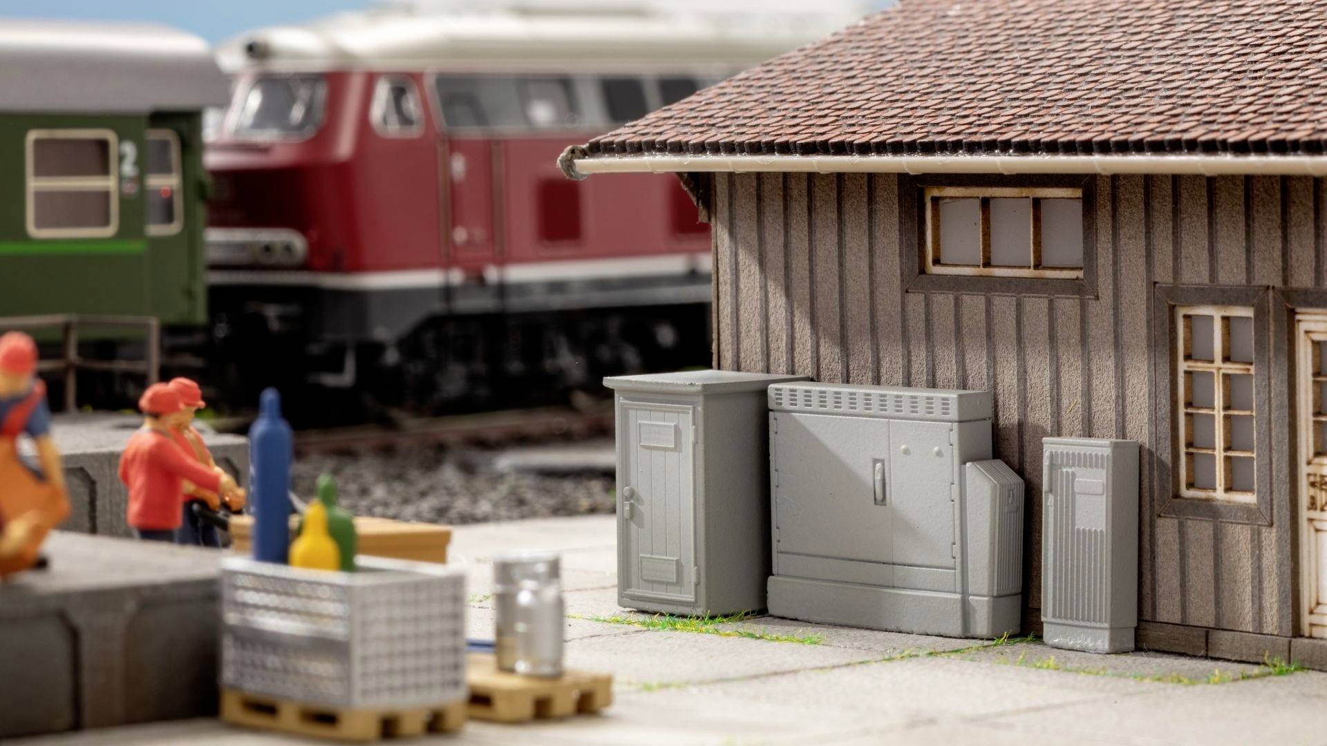 Miniature railway scene with a train, red train carriage in the background, two worker figures in orange clothing and a building in the foreground.