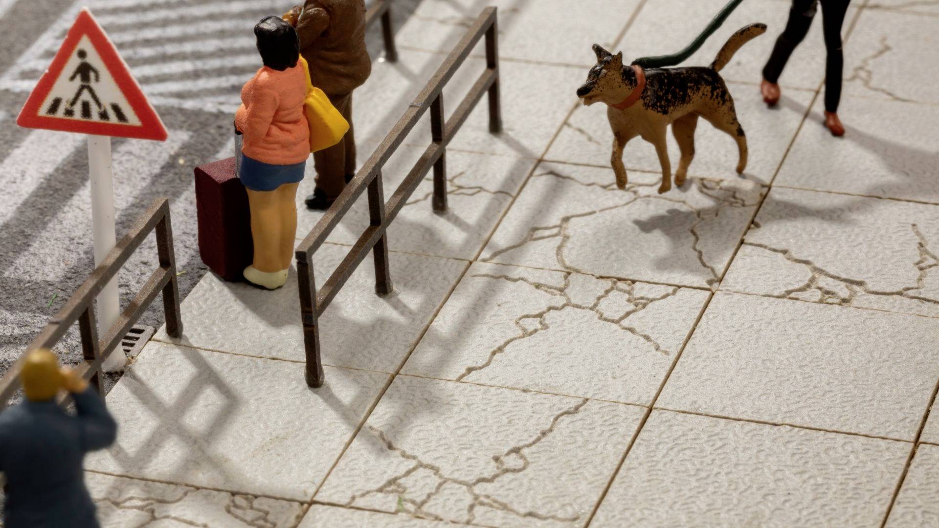 Several figures are standing on a pavement, a traffic light shows a pedestrian crossing sign, a dog is being walked on a lead.