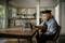 A person is sitting at a table with a guitar and looking at a smartphone propped up in front of a laptop. The room is comfortably furnished.