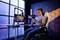 A woman is sitting in a gaming chair in front of a PC with two monitors and a webcam, playing a video game in a modern room.