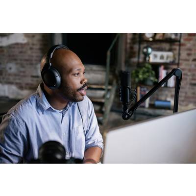 A man wearing headphones speaks into a microphone in a studio with brick walls, seated in front of a computer.