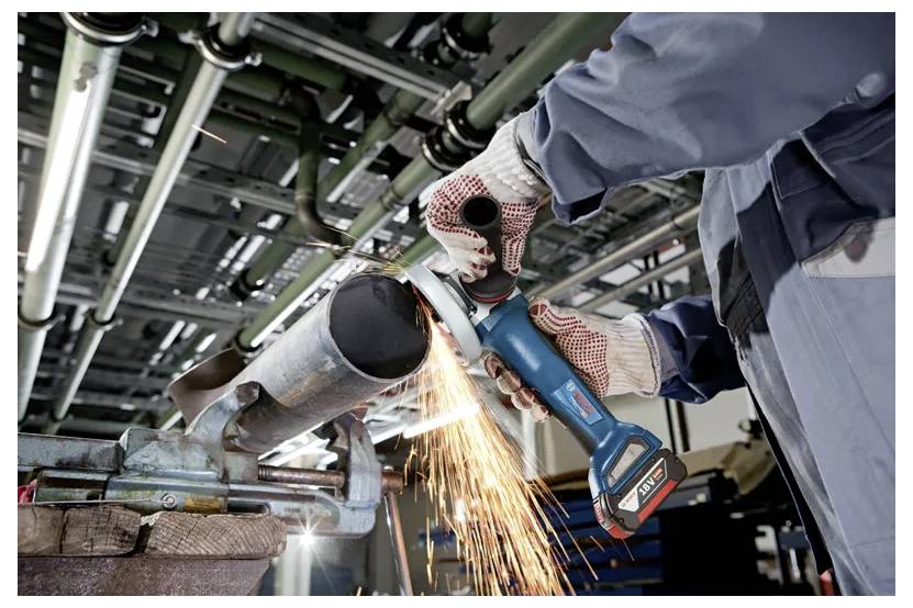A worker wearing gloves uses an angle grinder to cut a metal pipe, producing sparks in an industrial setting with pipes above.