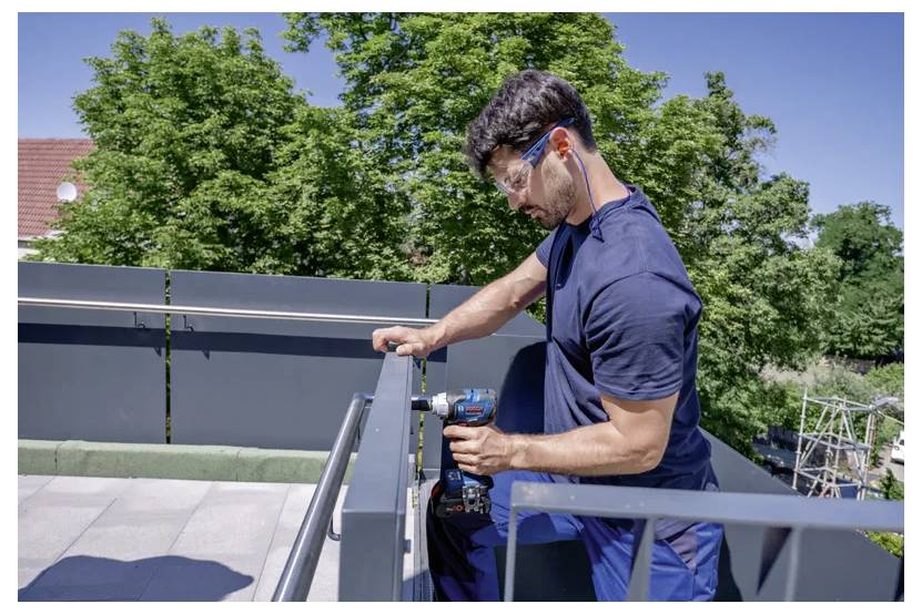 A person on a rooftop balcony using a power drill to fix a metal railing, with trees in the background.