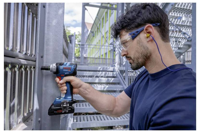 A man in safety gear uses an electric drill on a metal structure outdoors. He wears glasses and ear protection for safety.