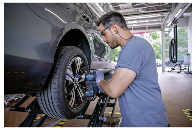 A mechanic uses a power tool to work on a car's wheel in an auto repair shop. He wears ear protection and gloves, focusing intently.
