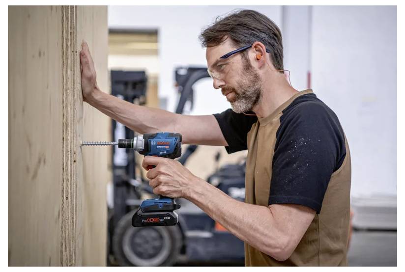A person in a workshop uses a blue cordless drill to drive a long screw into a wooden plank, with a forklift visible in the background.