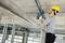 A worker wearing a hard hat and using a drill is securing a metal frame to the ceiling in an unfinished building.