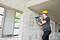 A construction worker wearing a yellow hard hat is drilling into a concrete wall with a hammer drill in an unfinished building.