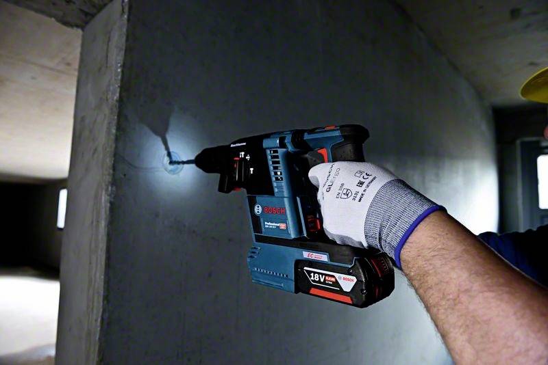 A person is drilling a hole in a concrete wall using a blue cordless hammer drill. They are wearing work gloves.
