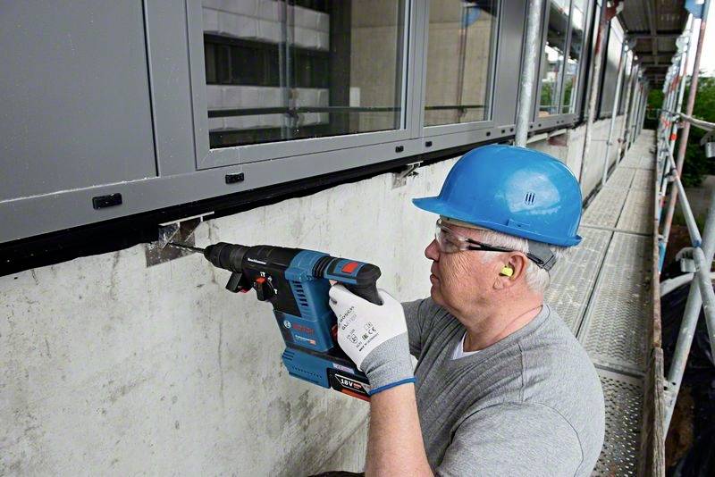 A worker wearing a hard hat and gloves is securing a metal construction to a concrete wall using an electric drill hammer.